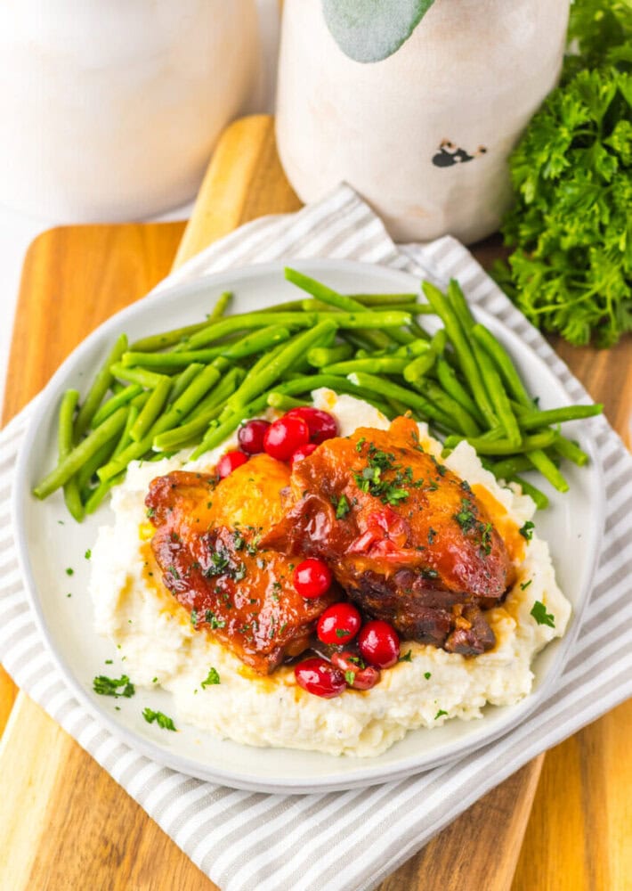 A plate with mashed potatoes topped with braised meat and cranberries, served with green beans, on a striped napkin and wooden board.
