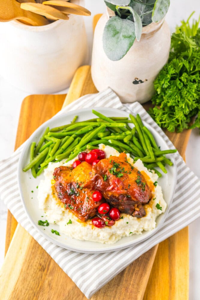 A plate of mashed potatoes topped with glazed chicken and cranberries, served with a side of green beans. The plate is on a striped napkin over a wooden board.