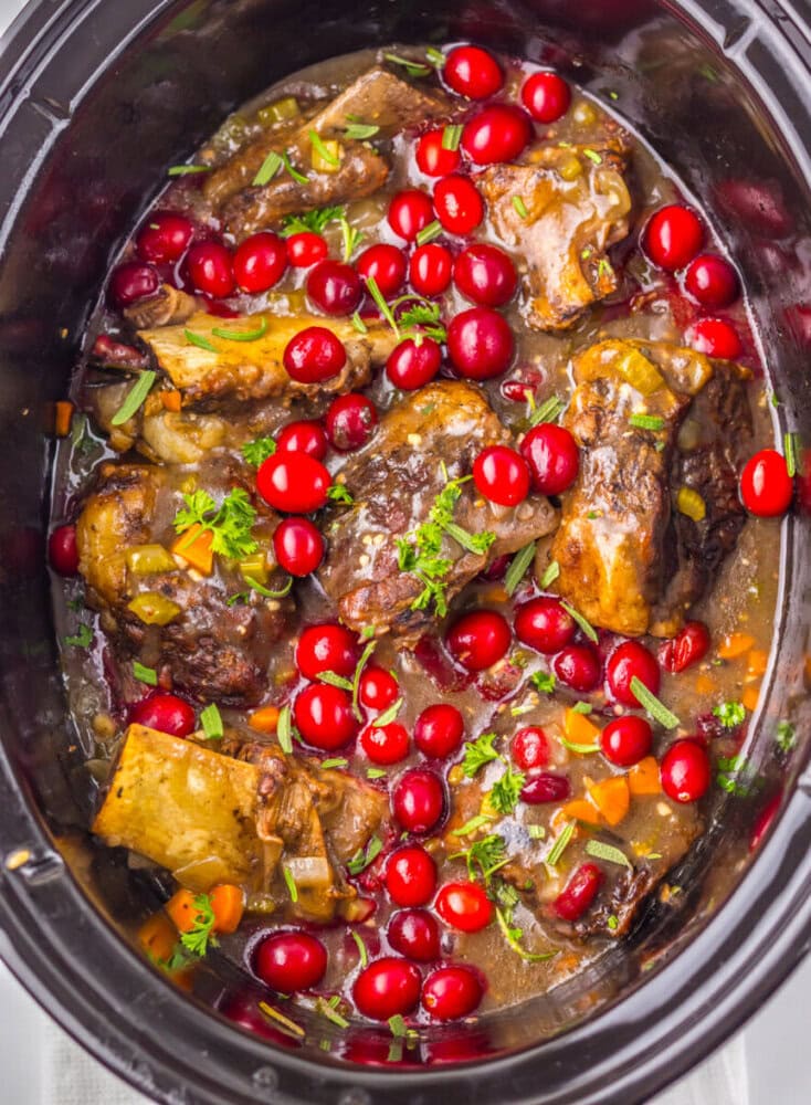Slow cooker filled with braised beef short ribs, garnished with fresh cranberries, chopped herbs, and vegetables in a rich sauce.