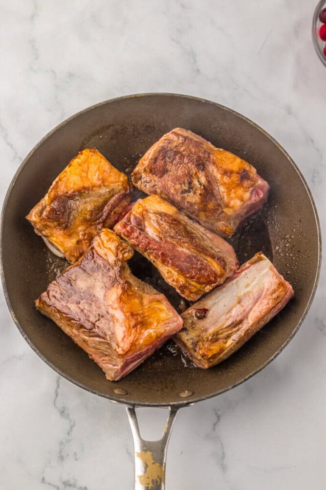 Five browned short ribs are searing in a black frying pan on a marble countertop.