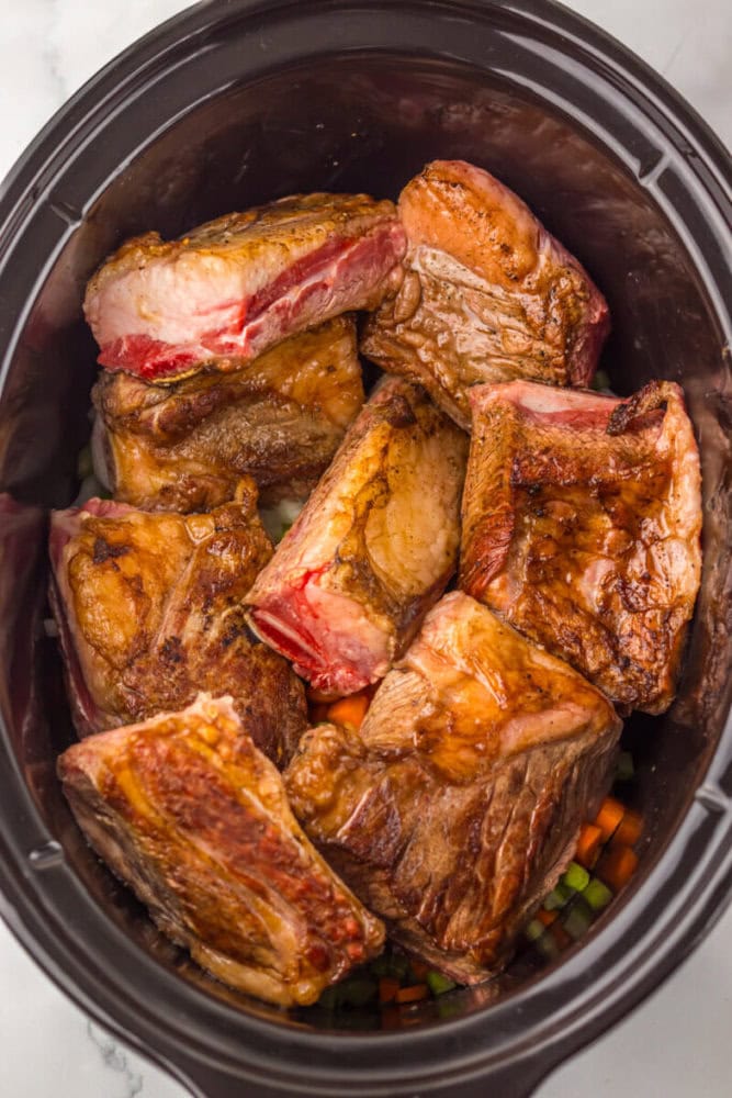 Browned beef short ribs arranged in a slow cooker, placed over a layer of chopped carrots and celery.