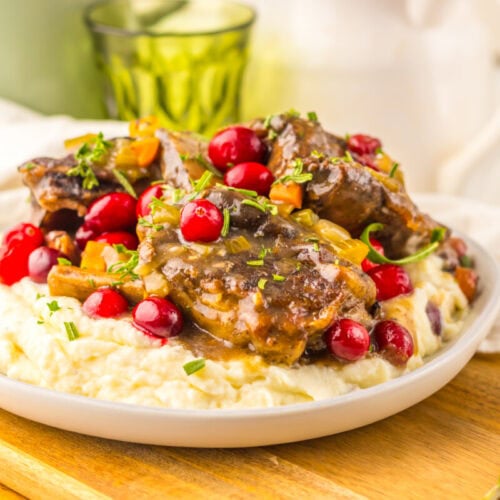 A plate of mashed potatoes topped with braised meat, cranberries, chopped herbs, and vegetables, served on a wooden board.