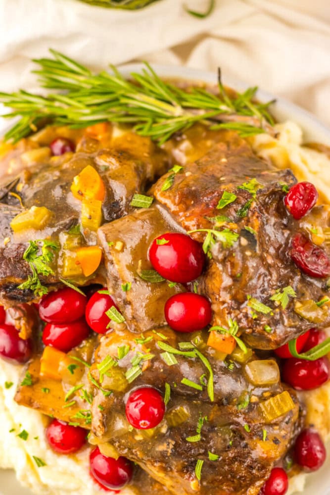 Braised beef short ribs with gravy, garnished with fresh cranberries, chopped herbs, and a sprig of rosemary, served on a bed of mashed potatoes.