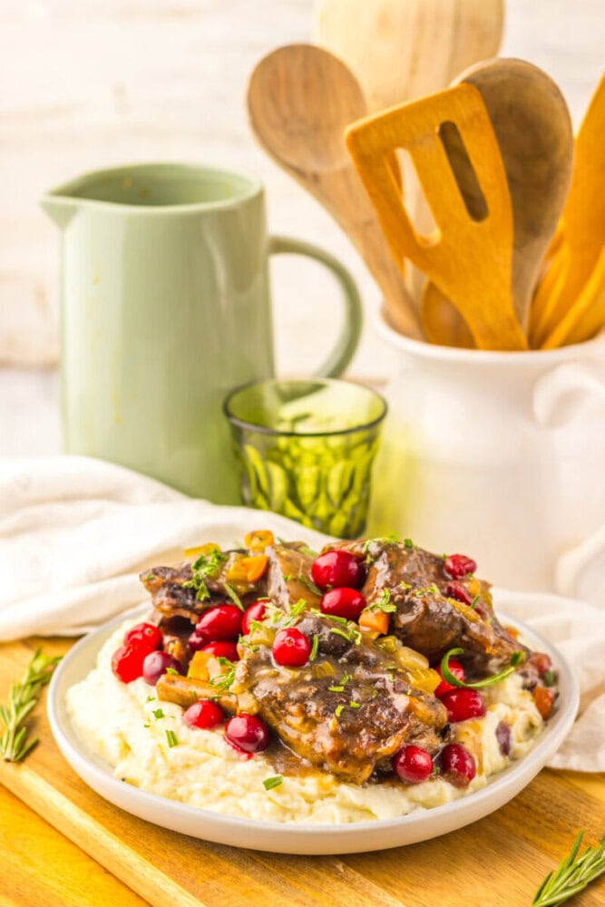 A plate of braised meat with cranberries and chopped herbs is served on mashed potatoes, with kitchen utensils and a green pitcher in the background.