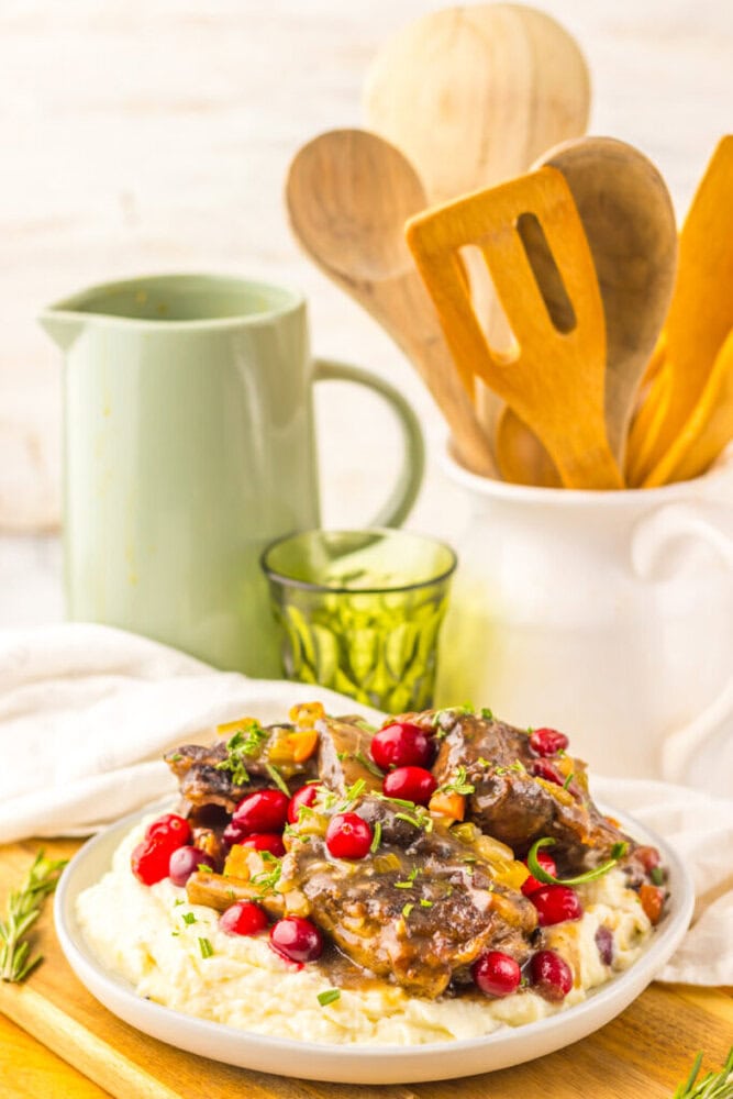 A plate of braised meat with gravy sits on mashed potatoes, garnished with cranberries and herbs. A green pitcher, glass, and wooden utensils are in the background.
