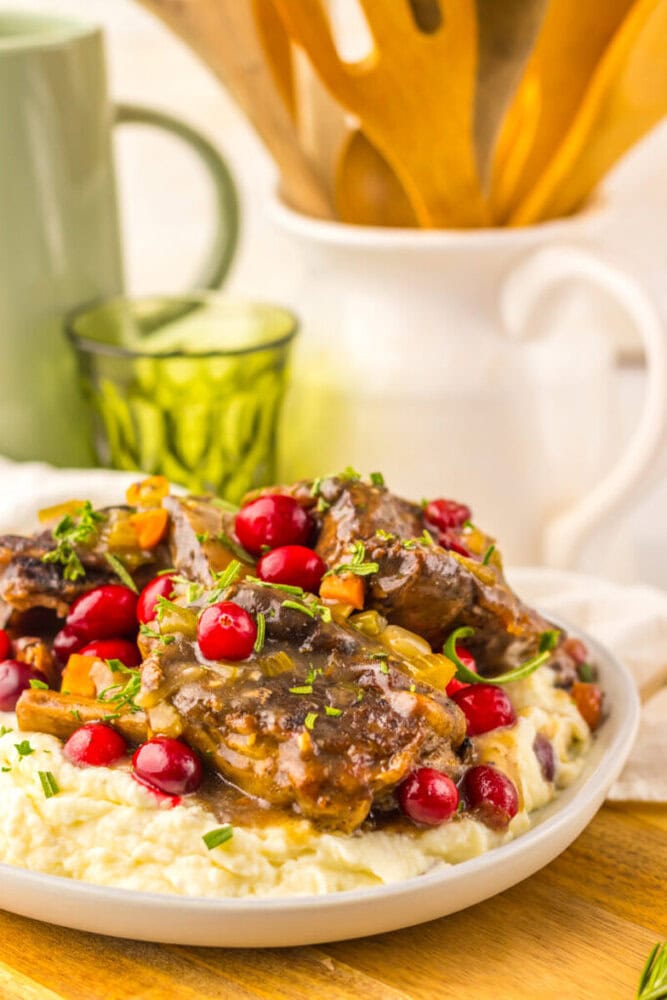 Braised meat with vegetables and cranberries served over mashed potatoes on a white plate, with a green glass and kitchen utensils in the background.