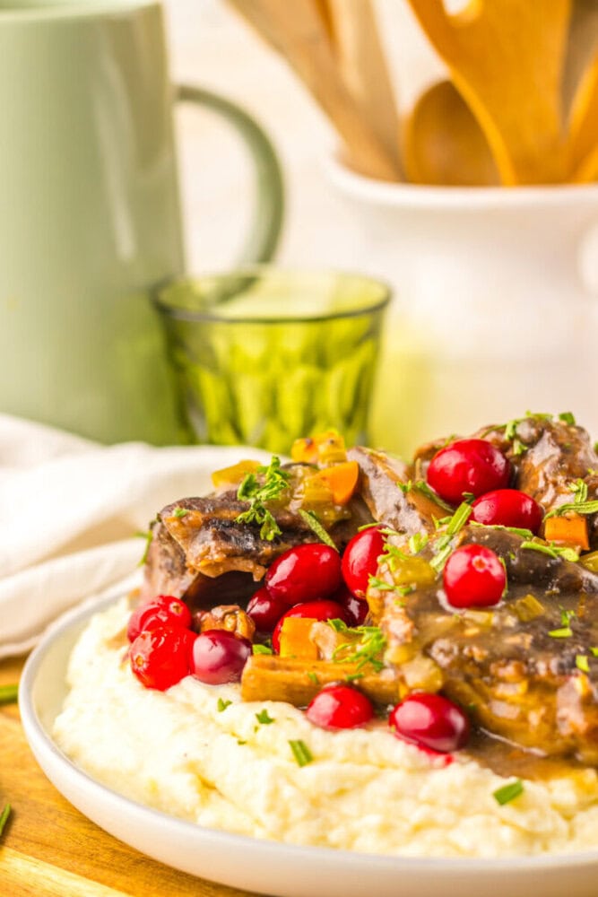 A plate of creamy mashed potatoes topped with braised meat, cranberries, chopped vegetables, and fresh herbs sits on a wooden surface. A green mug and glass are in the background.