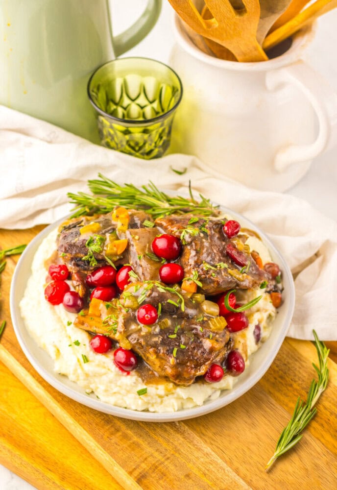 A plate of braised meat with a brown sauce, garnished with fresh cranberries and herbs, served on a bed of mashed potatoes. Wooden utensils and kitchenware are in the background.