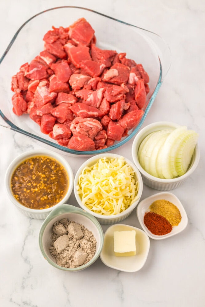 A glass bowl of raw beef chunks surrounded by sliced onions, shredded cheese, butter, gravy mix, spices, and a bowl of sauce on a marble surface.