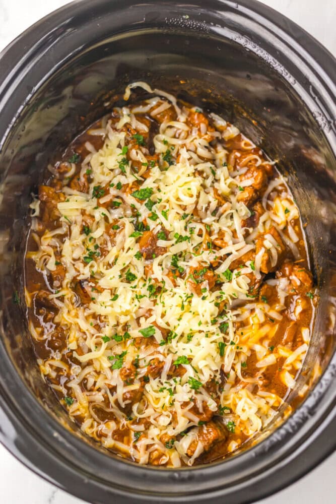 Slow cooker filled with beef stew topped with shredded cheese and chopped parsley.