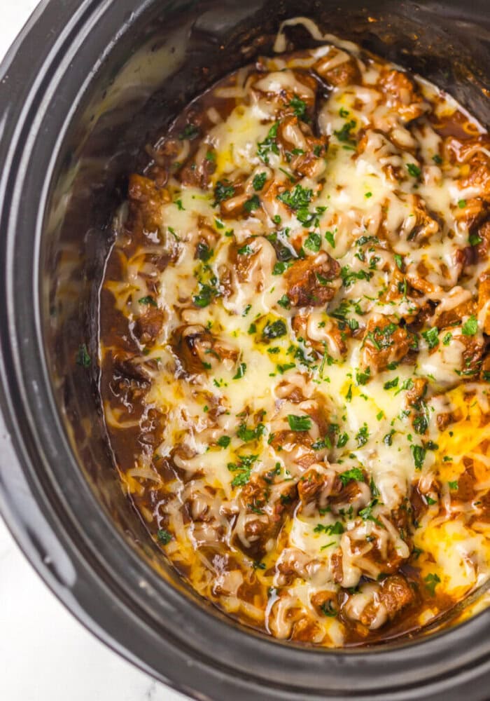 Shredded beef topped with melted cheese and chopped herbs cooked in a black slow cooker.