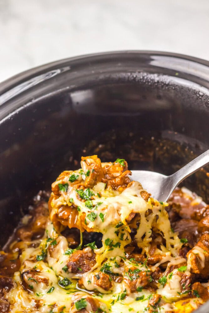 A spoon lifts cheesy, cooked beef with herbs from a black slow cooker.