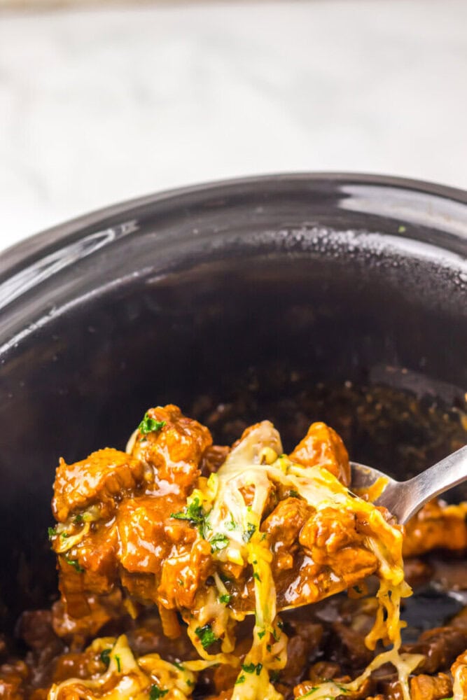 A fork lifts a serving of cheesy, saucy beef from a black slow cooker.