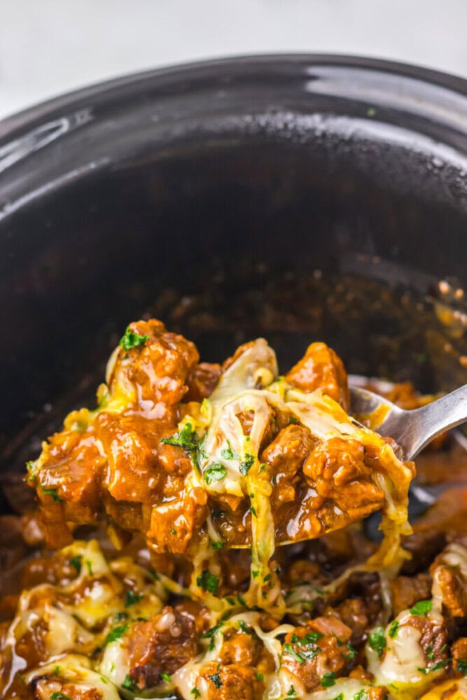 A close-up of a spoon lifting a cheesy, saucy beef dish from a black slow cooker.
