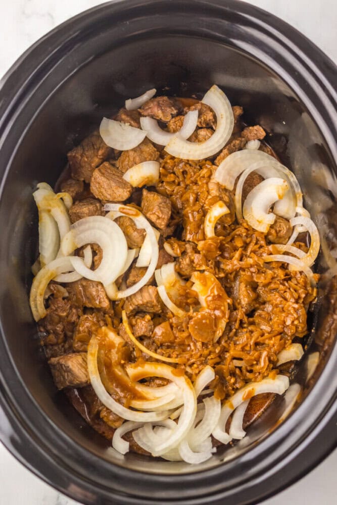 A slow cooker filled with cooked beef chunks, sliced onions, and a brown sauce or seasoning mixture.