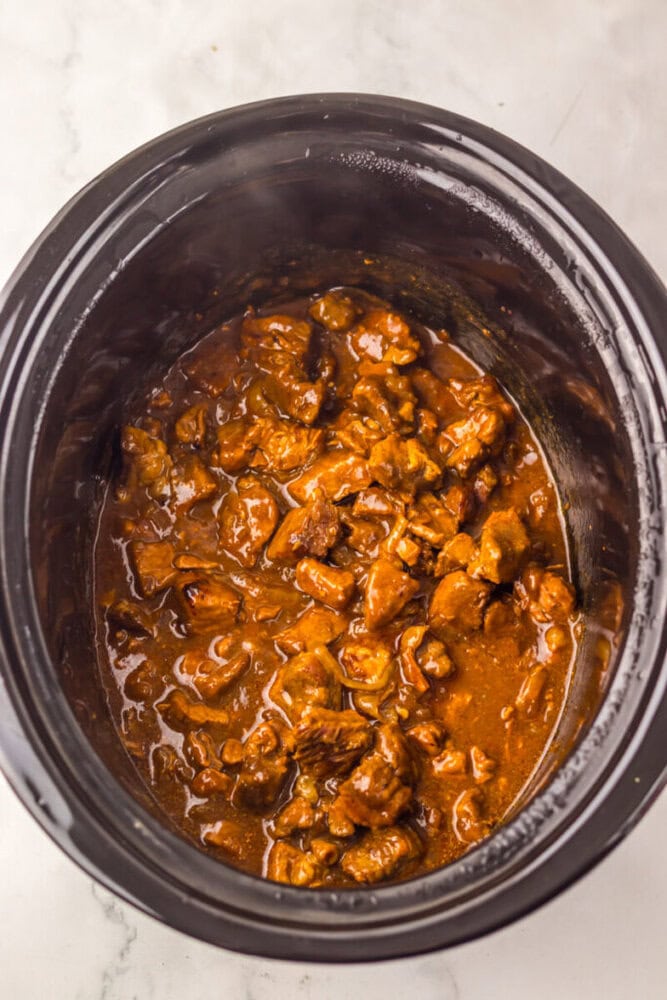 Chunks of beef in a rich brown sauce cook inside a black slow cooker on a white surface.
