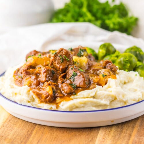 A plate of mashed potatoes topped with beef stew and garnished with Brussels sprouts, served on a wooden board.