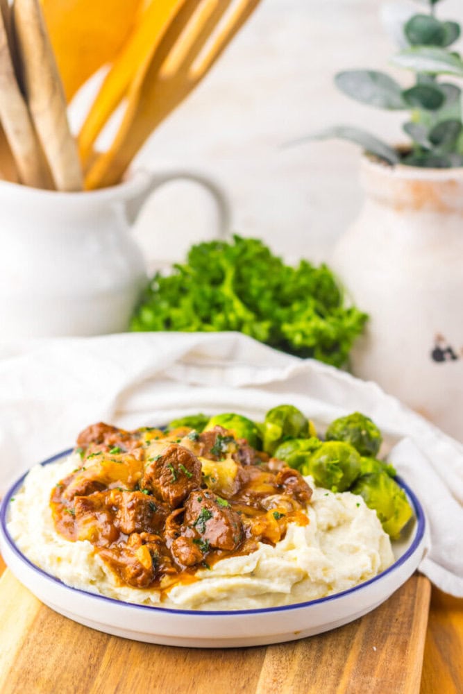 A plate of mashed potatoes topped with beef stew and accompanied by Brussels sprouts, set on a wooden board with kitchen utensils, greens, and a white pitcher in the background.