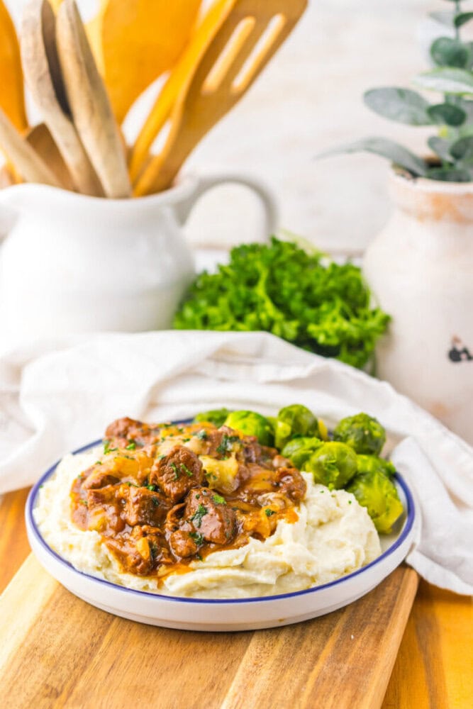 Plate of mashed potatoes topped with beef stew, served with Brussels sprouts on the side, on a wooden board with kitchen utensils and greenery in the background.