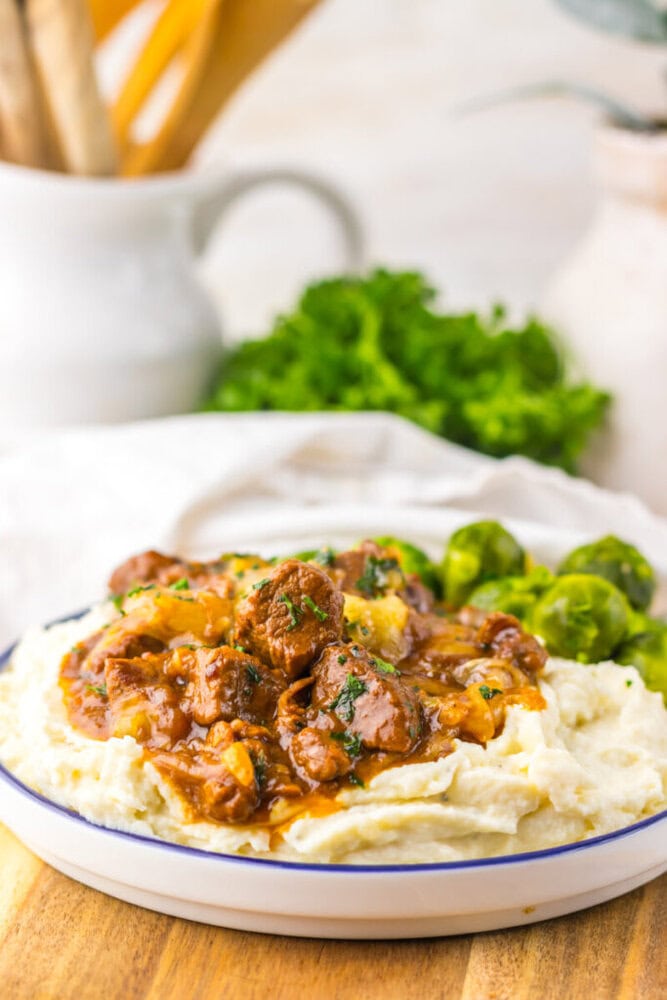Plate of mashed potatoes topped with beef stew and gravy, served with Brussels sprouts on the side.