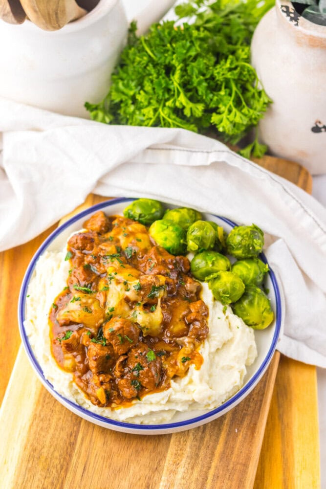 A bowl of mashed potatoes topped with beef stew and served with Brussels sprouts on a wooden board, with fresh parsley in the background.