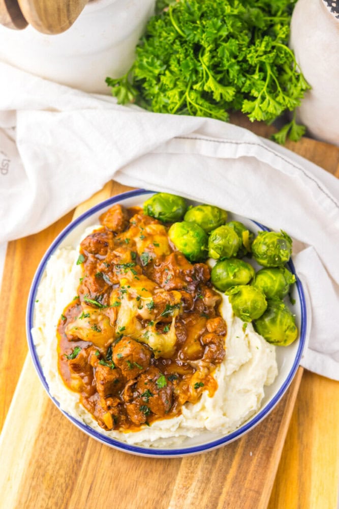 A plate of mashed potatoes topped with beef stew and a side of Brussels sprouts, set on a wooden board with fresh parsley in the background.