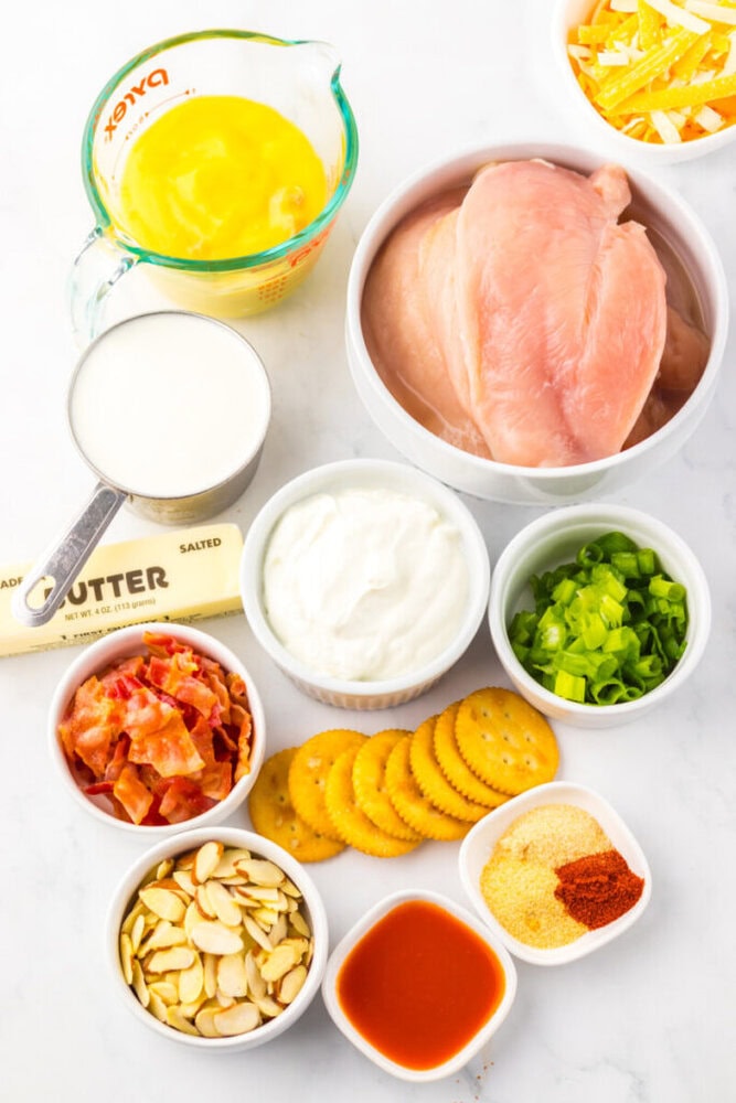 Assorted casserole ingredients on a white surface, including raw chicken breasts, eggs, milk, yogurt, green onions, bacon, crackers, sliced almonds, butter, hot sauce, and seasonings.