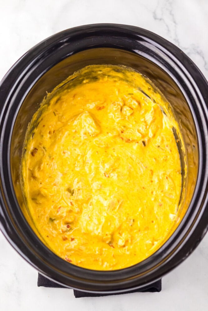 A slow cooker filled with creamy, yellow cheese dip with visible chunks of peppers and spices on a marble surface.