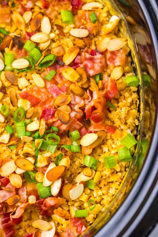 Close-up of a cooked dish topped with sliced almonds, chopped green onions, and pieces of bacon in a black slow cooker.