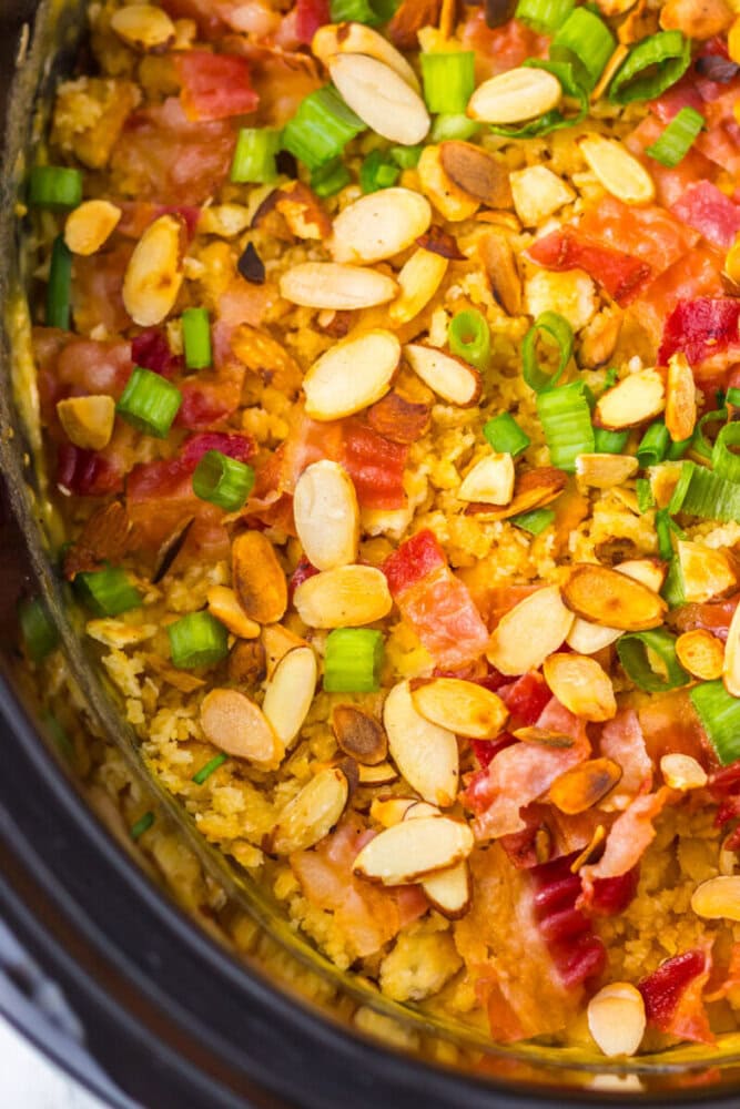 A close-up of a casserole topped with sliced almonds, chopped green onions, and pieces of bacon in a slow cooker.