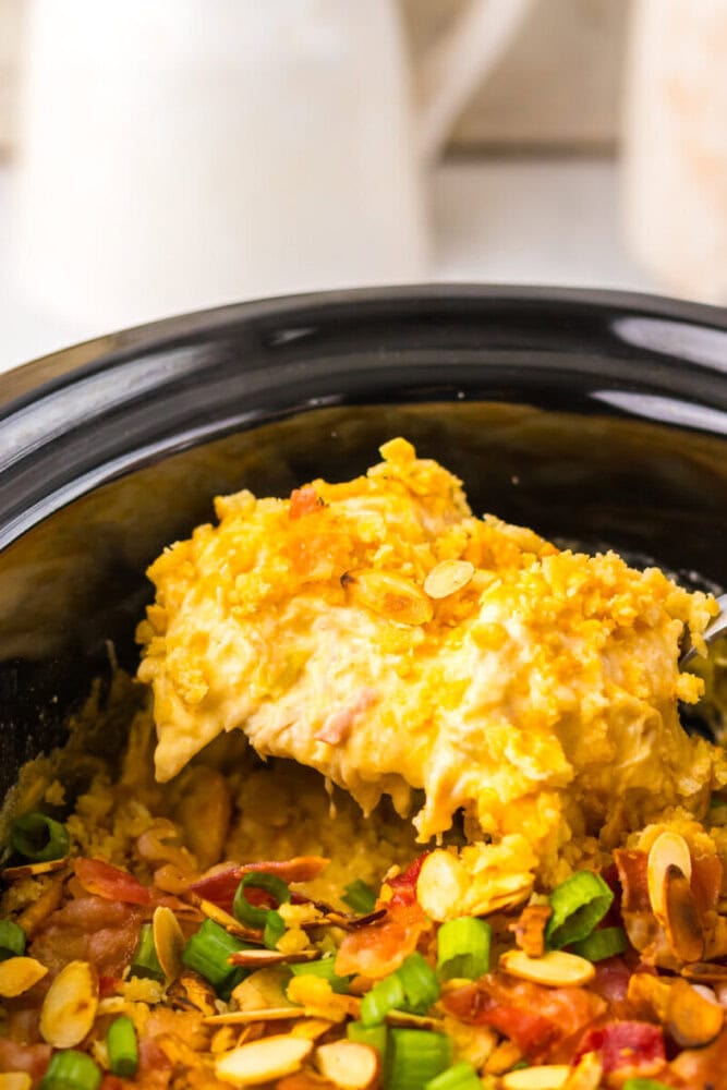 A serving of creamy casserole with a crunchy topping is being scooped from a black slow cooker; sliced green onions and almonds are visible on top.