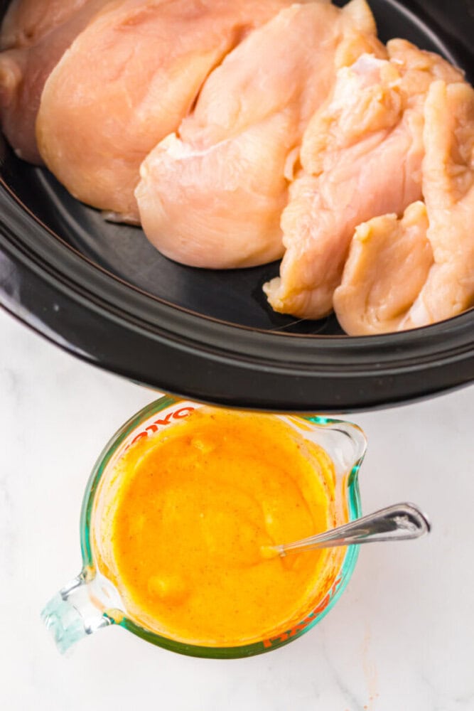 Raw chicken breasts in a slow cooker next to a measuring cup filled with yellow-orange sauce and a spoon.