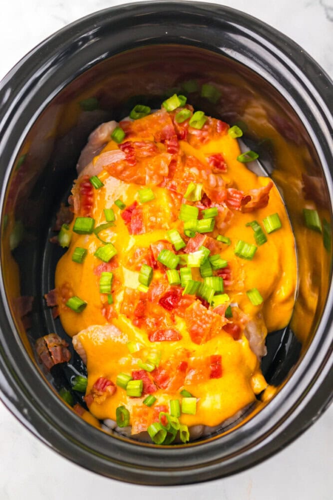 Chicken breasts in a slow cooker topped with melted cheddar cheese, chopped bacon, and sliced green onions.
