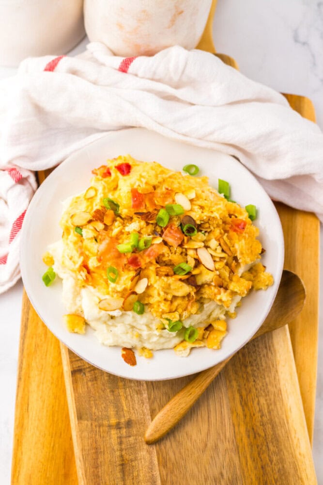 A plate of creamy mashed potatoes topped with a cheesy chicken casserole, chopped green onions, and almond slices, placed on a wooden board with a wooden spoon nearby.