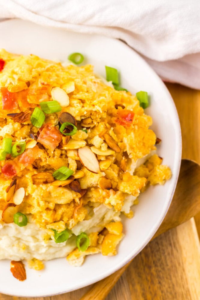 A close-up of a mashed potato casserole topped with crushed crackers, sliced almonds, bacon pieces, and chopped green onions on a white plate.