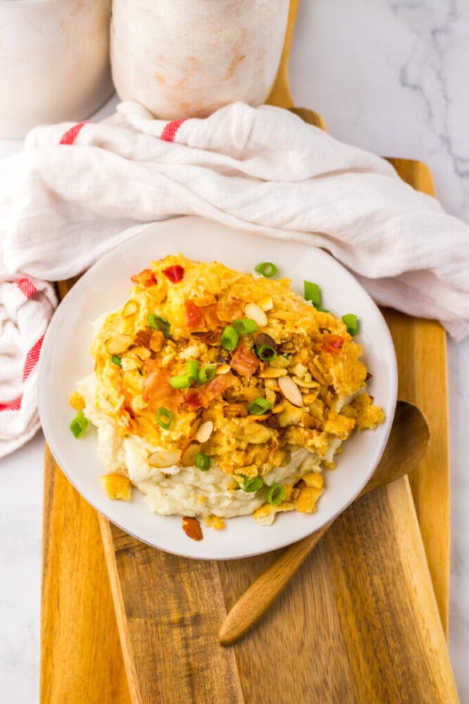 A plate of scrambled eggs with chopped green onions and sliced almonds served over mashed potatoes, placed on a wooden board with a wooden spoon nearby.