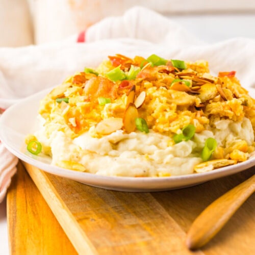 A plate of mashed potatoes topped with scrambled eggs, sliced almonds, green onions, and crispy bacon on a wooden board with a wooden spoon beside it.