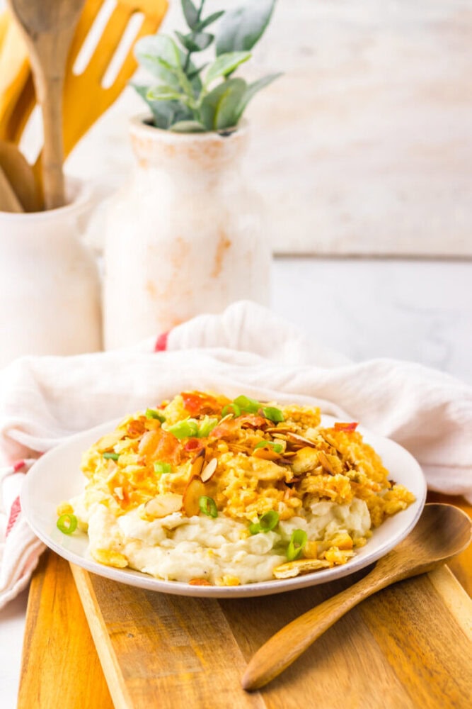 A plate of creamy casserole topped with sliced almonds and chopped green onions, placed on a wooden board with a wooden spoon beside it.