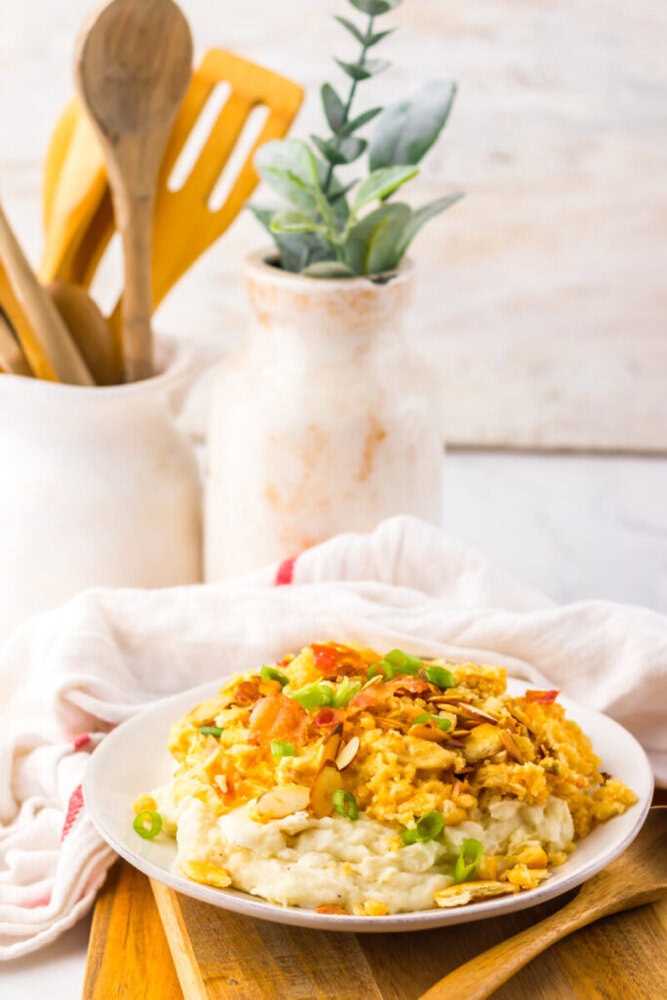 A plate of mashed potatoes topped with a yellow scrambled mixture, green onions, and sliced almonds, set on a wooden board with utensils and a decorative plant in the background.