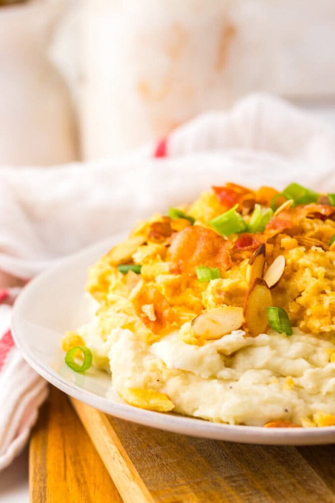 A plate of mashed potatoes topped with a creamy casserole, sliced almonds, chopped green onions, and crispy bacon bits.