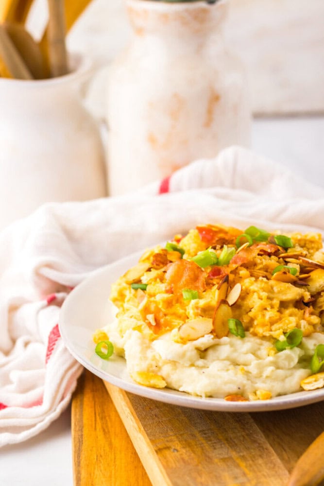 A plate of scrambled eggs topped with sliced almonds and green onions, served over mashed potatoes on a white plate placed on a wooden board.