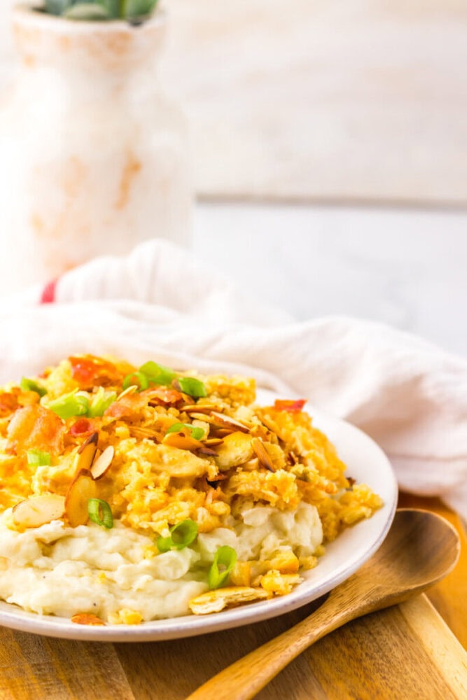 A white plate with mashed potatoes topped with a yellow casserole, garnished with sliced almonds and green onions, on a wooden board with a spoon and a white napkin nearby.