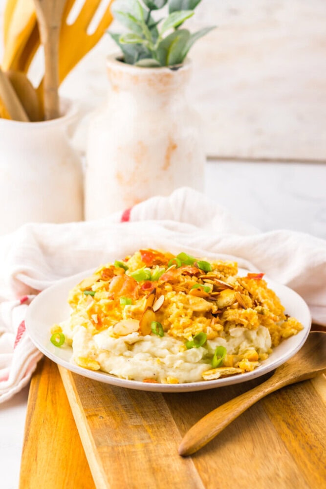 A plate of creamy rice topped with scrambled eggs, chopped green onions, sliced almonds, and diced tomatoes on a wooden board, with kitchen utensils and a potted plant in the background.