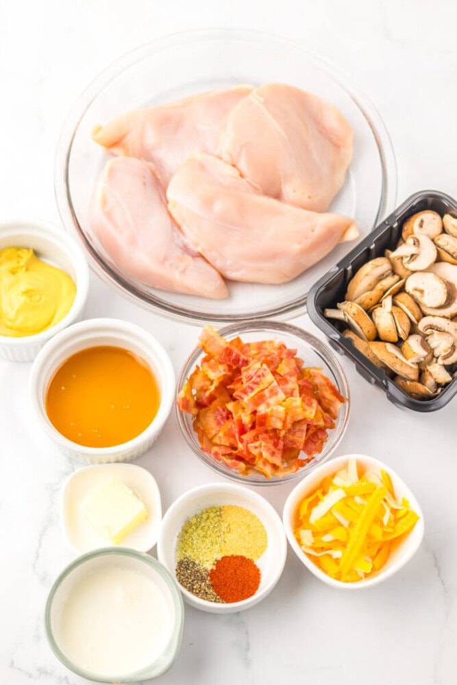 A bowl of raw chicken breasts, a container of sliced mushrooms, chopped cooked bacon, mustard, honey, butter, cream, sliced yellow peppers, and various spices in small bowls.