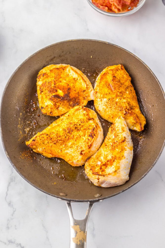 Four seasoned chicken breasts cooking in a nonstick skillet on a white marble countertop.