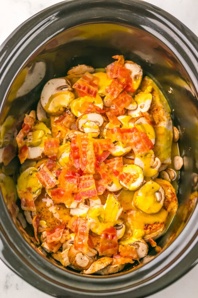 Sliced mushrooms, cooked bacon pieces, and melted cheese over seasoned chicken breasts in a slow cooker.