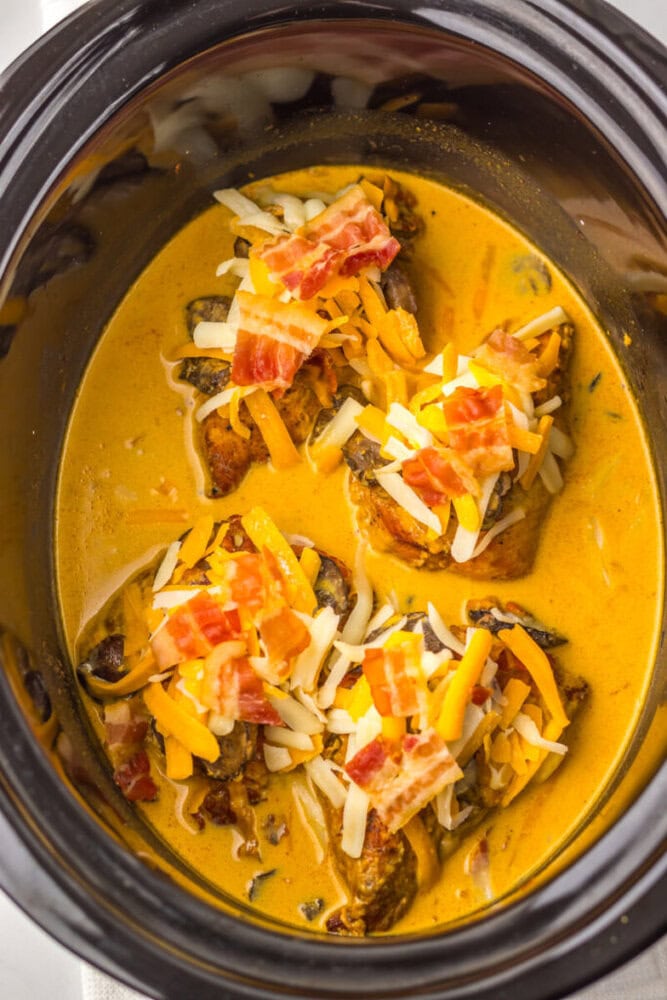 Chicken breasts in a creamy orange sauce topped with shredded cheese and bacon, inside a slow cooker.