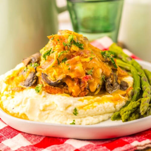 A plate of mashed potatoes topped with a creamy chicken and mushroom mixture, garnished with herbs, and served with asparagus on the side.
