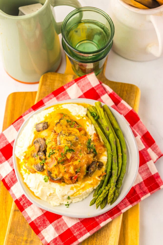 Plate of mashed potatoes topped with a mushroom and gravy mixture, served with steamed asparagus on the side, placed on a red-and-white checkered napkin.