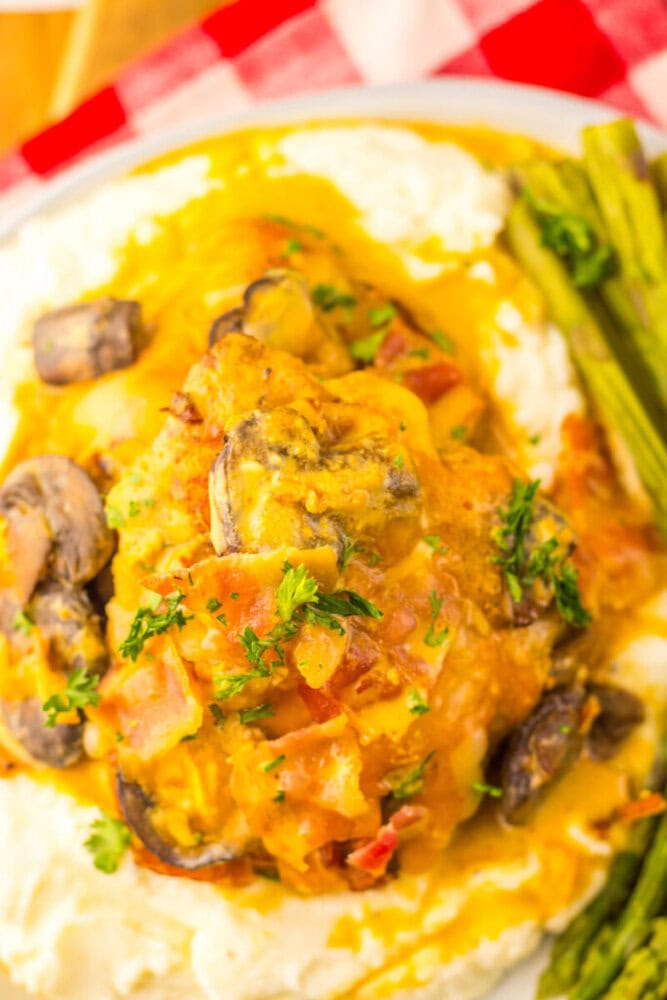 A plate with creamy mashed potatoes topped with chicken, sautéed mushrooms, and sauce, garnished with parsley, served with a side of asparagus.